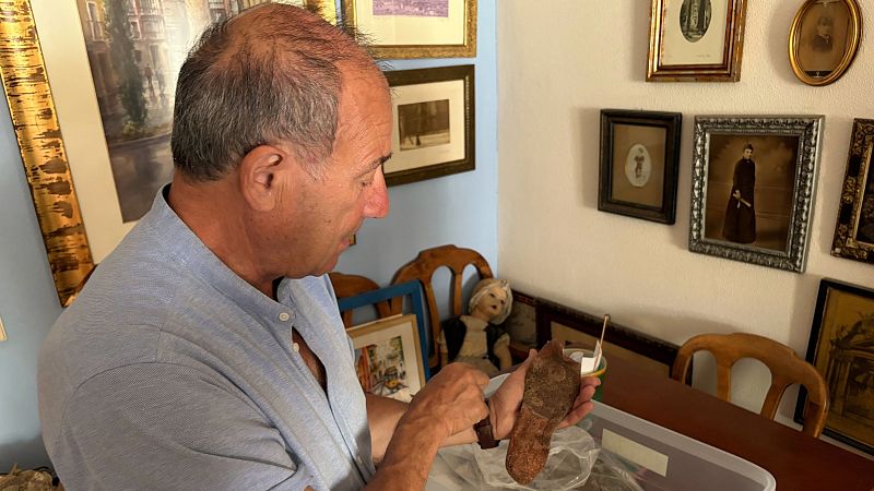 Julio del Olmo sostiene una suela de zapato de víctima del franquismo, rodeado de cuadros y objetos antiguos en un interior.
