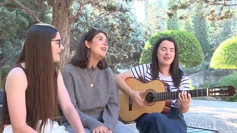 Tres mujeres jóvenes en un parque soleado, una tocando la guitarra acústica y las otras dos cantando o escuchando, con exuberante vegetación de fondo.