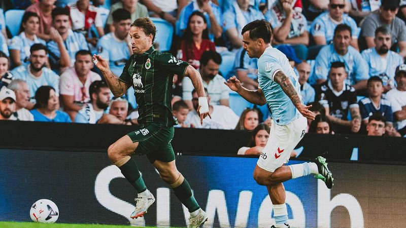 Iñigo Vicente del Racing y Iago Aspas del Celta compiten por el balón en un partido de fútbol, con el estadio lleno de espectadores al fondo.