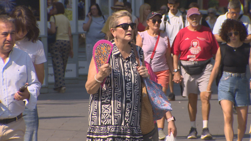 Mujer con vestido estampado y abanico, rodeada de gente en un día soleado y caluroso. Se aprecian gafas de sol y vestimenta ligera.