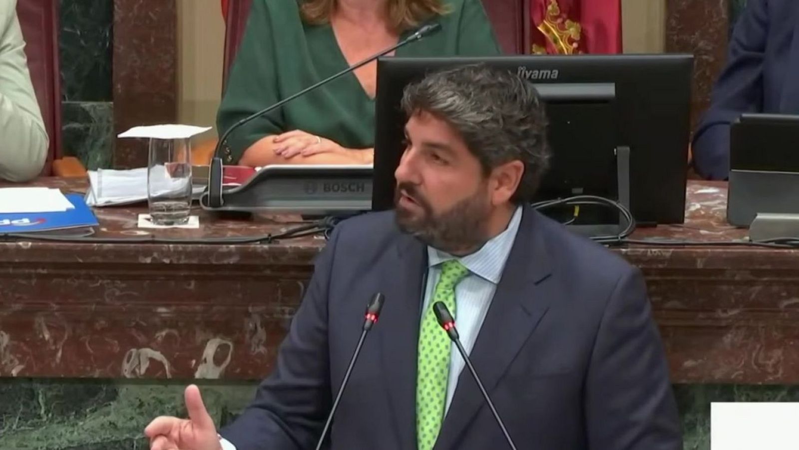 Fernando López Miras, Presidente de la Región de Murcia, habla en la Asamblea Regional de Murcia. Lleva traje azul, camisa azul y corbata verde lima. En la mesa, micrófonos, vaso de agua y carpeta del PP. Se aprecian logos de Iiyama y Bosch.