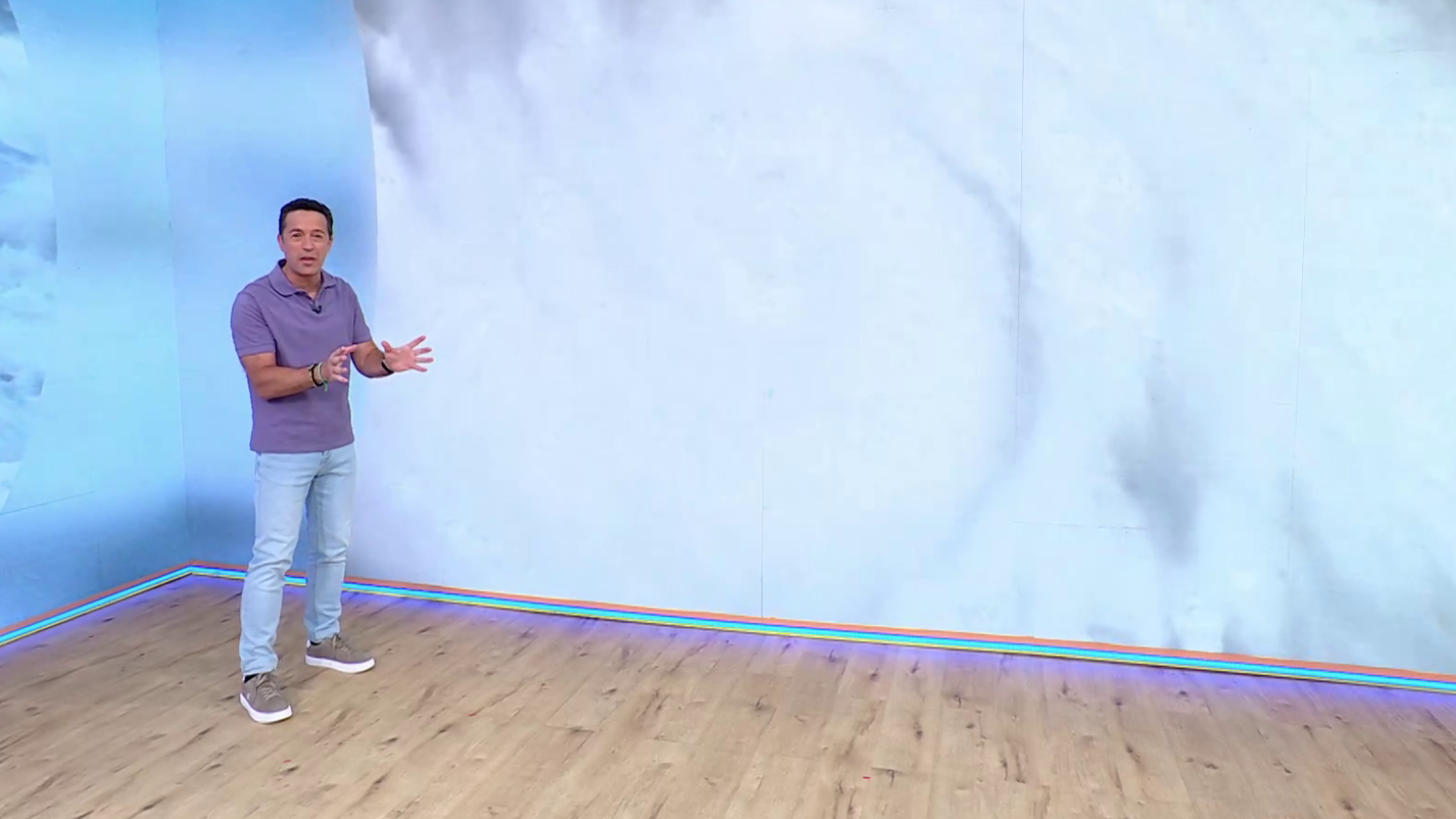 Roberto Brasero, presentador de televisión, posa en un plató con un fondo digital abstracto de tonos azules y blancos. Lleva una camiseta polo morada y jeans, con las manos abiertas en un gesto explicativo.