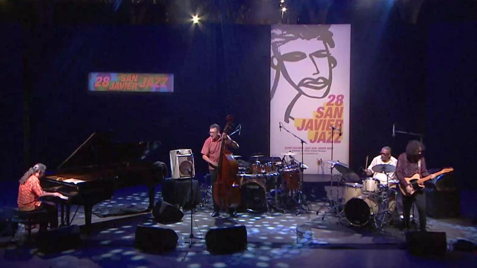 Concierto de jazz con cuatro músicos en vivo. Se anuncia "28 SAN JAVIER JAZZ" del 1 al 26 de julio de 2015. Piano, contrabajo, batería y guitarra eléctrica en escena.