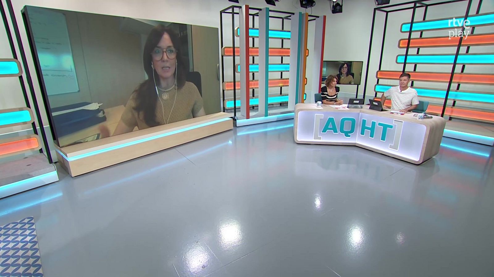Plató de televisión con presentadores Marta Cillán y Jon Ariztimuño, escritorio "AQHT" y pantalla con participante remota. Fondo con luces azules/celestes y naranjas/rojizas, logo "rtve play".