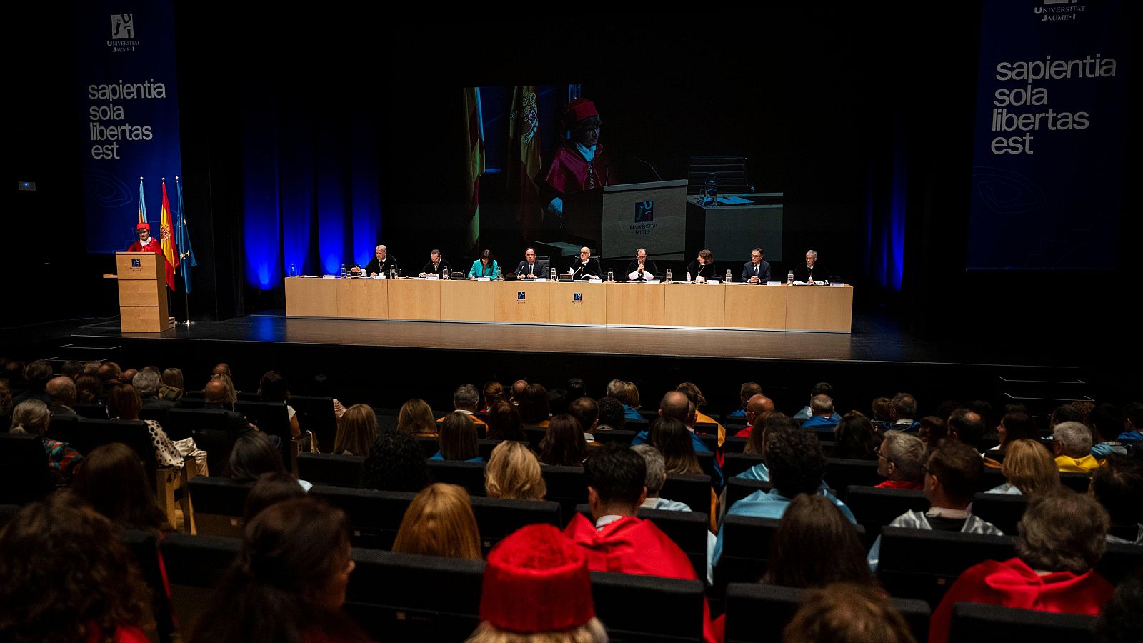 Orador con toga y birrete rojo en atril, flanqueado por banderas de España y UH. Mesa presidencial y audiencia atenta en auditorio.