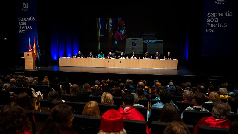 Orador con toga y birrete rojo en atril, flanqueado por banderas de España y UH. Mesa presidencial y audiencia atenta en auditorio.
