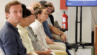 Grupo de personas atentas en una sala, mirando hacia la derecha. Se aprecian detalles de vestimenta, una pantalla con "ARCONTE" y un extintor rojo.