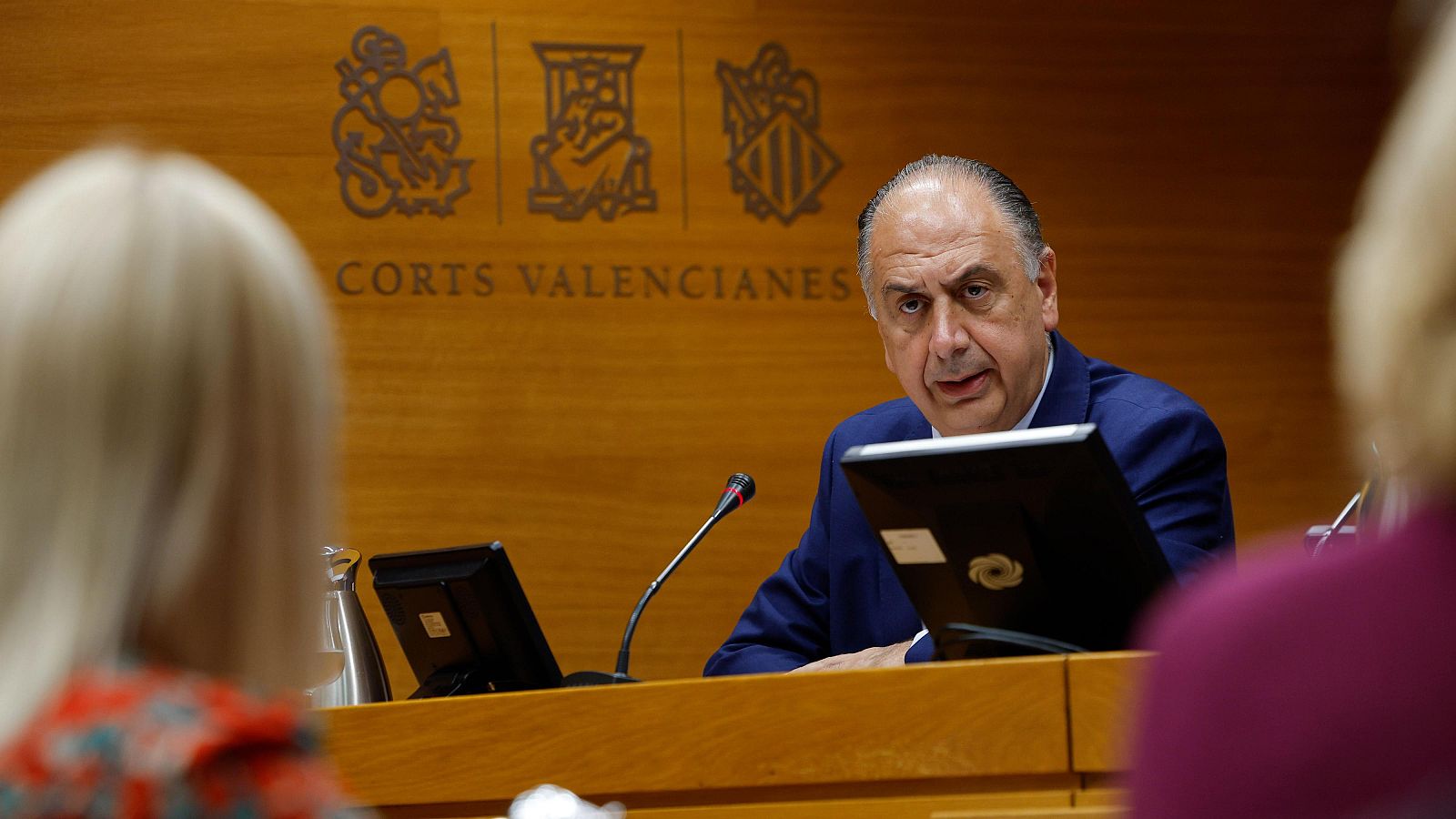 Ximo Puig, expresidente de la Generalitat Valenciana, en las Corts Valencianes, con traje azul, micrófono y monitores. Fondo con "CORTS VALENCIANES" y emblemas.