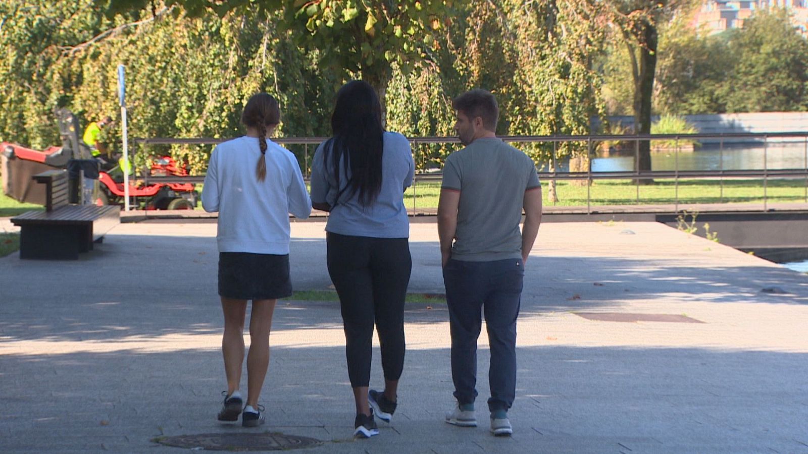 Tres personas, dos mujeres y un hombre, caminan por un sendero exterior con vegetación y agua. Se aprecian detalles de su vestimenta y calzado. Al fondo, una máquina cortacésped roja.