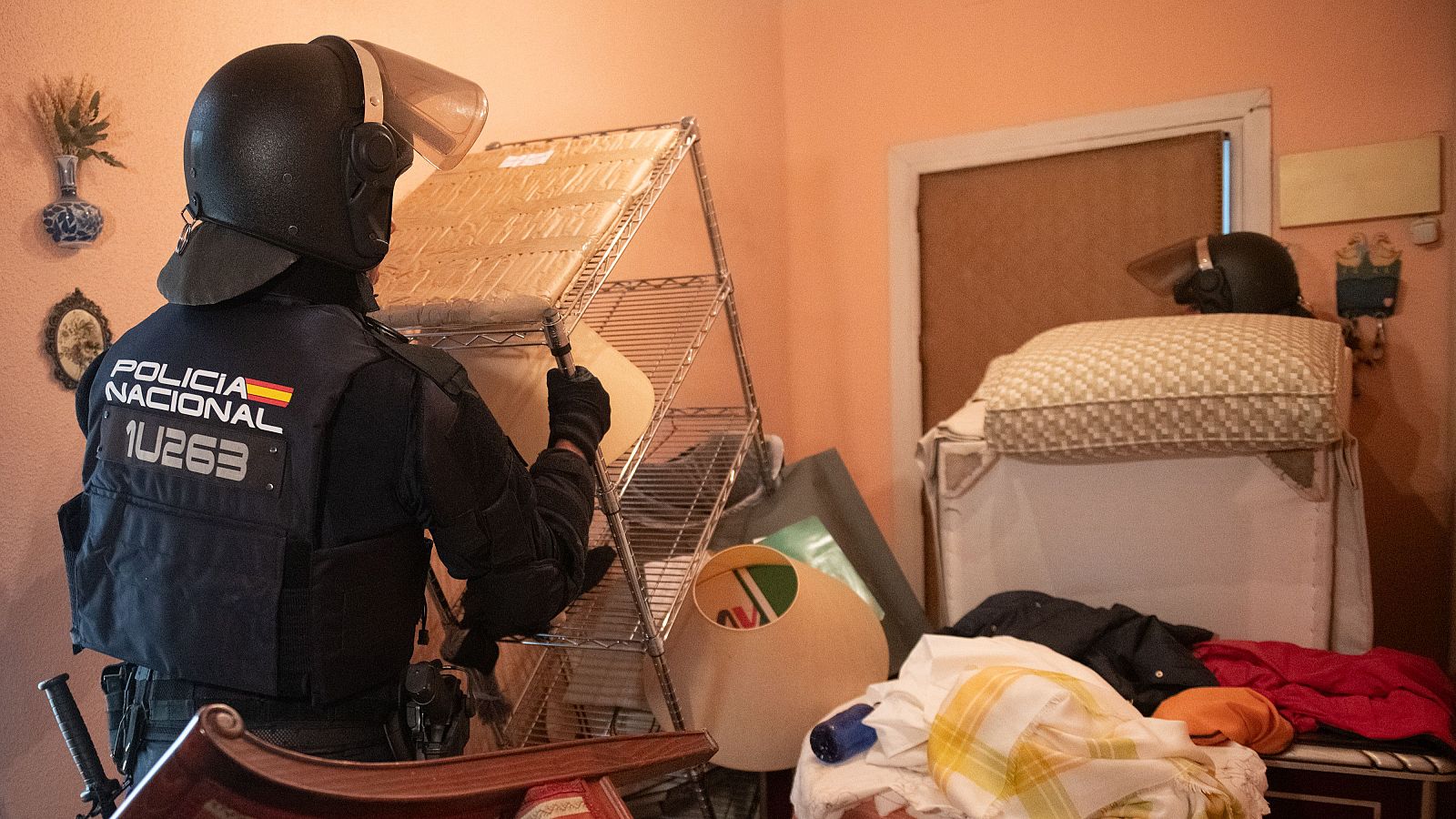 Agente de la Policía Nacional con chaleco táctico y casco antidisturbios, número 1U263, manipula un estante en una habitación desordenada. Se aprecian pertenencias y un televisor antiguo.