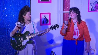 Mujeres en escenario con guitarra Epiphone verde, copas y cuadro "THIS GIRL IS ON FIRE".