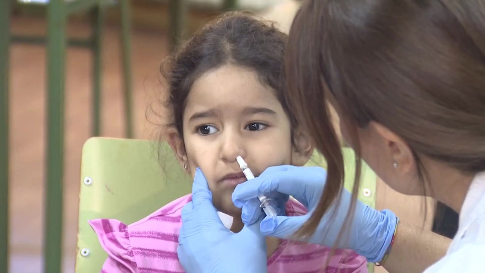 Niña escolar con camiseta a rayas rosas y blancas recibe vacuna en aerosol nasal de profesional sanitaria con bata blanca y guantes azules. La vacuna se administra en la fosa nasal derecha.