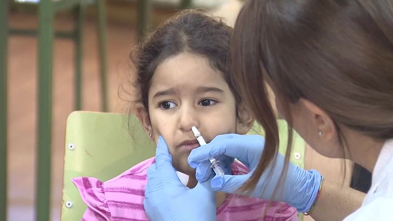 Niña escolar con camiseta a rayas rosas y blancas recibe vacuna en aerosol nasal de profesional sanitaria con bata blanca y guantes azules. La vacuna se administra en la fosa nasal derecha.