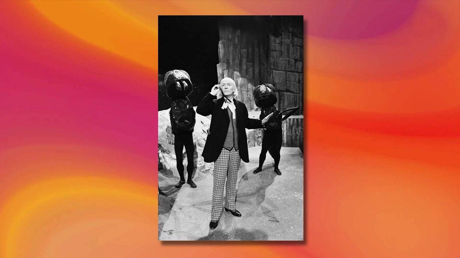 William Hartnell, el Primer Doctor, junto a dos "Monoids" en una producción de ciencia ficción en blanco y negro. El Doctor viste abrigo oscuro y chaleco claro.