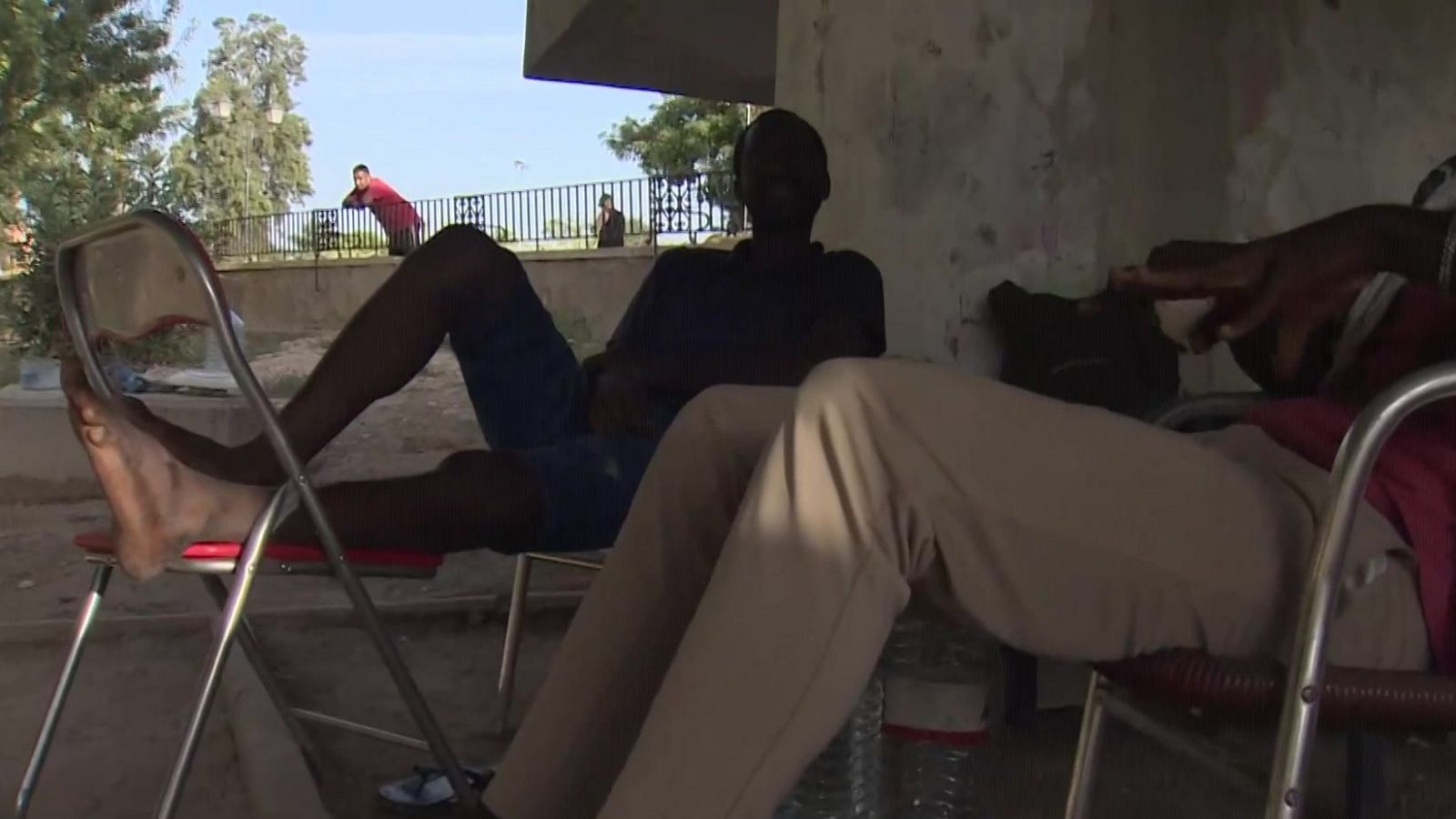 Hombres sentados en sillas plegables en sombra, con personas al sol detrás de una barandilla. Cielo azul y árboles al fondo.