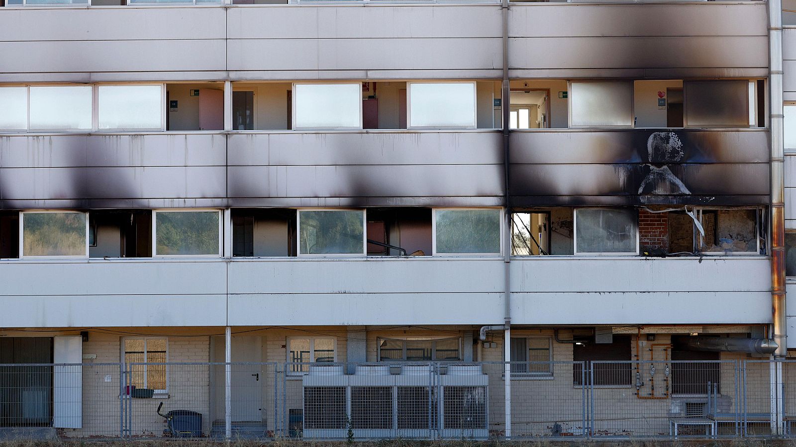 Fachada de edificio residencial con daños severos por incendio, hollín negro y ventanas rotas en plantas superiores. Planta baja intacta con valla y unidades exteriores.