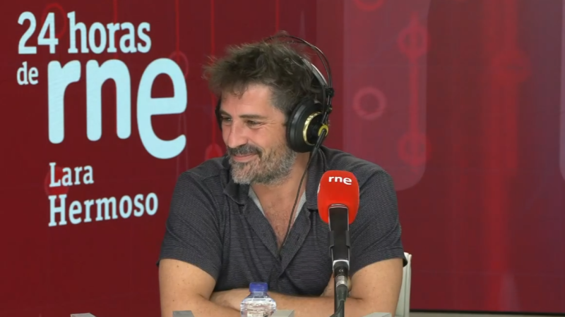Juan Gómez-Jurado en estudio de radio, con auriculares y camiseta gris. Micrófono rojo RNE y botella de agua. Fondo rojo con "24 horas de rne" y "Lara Hermoso".