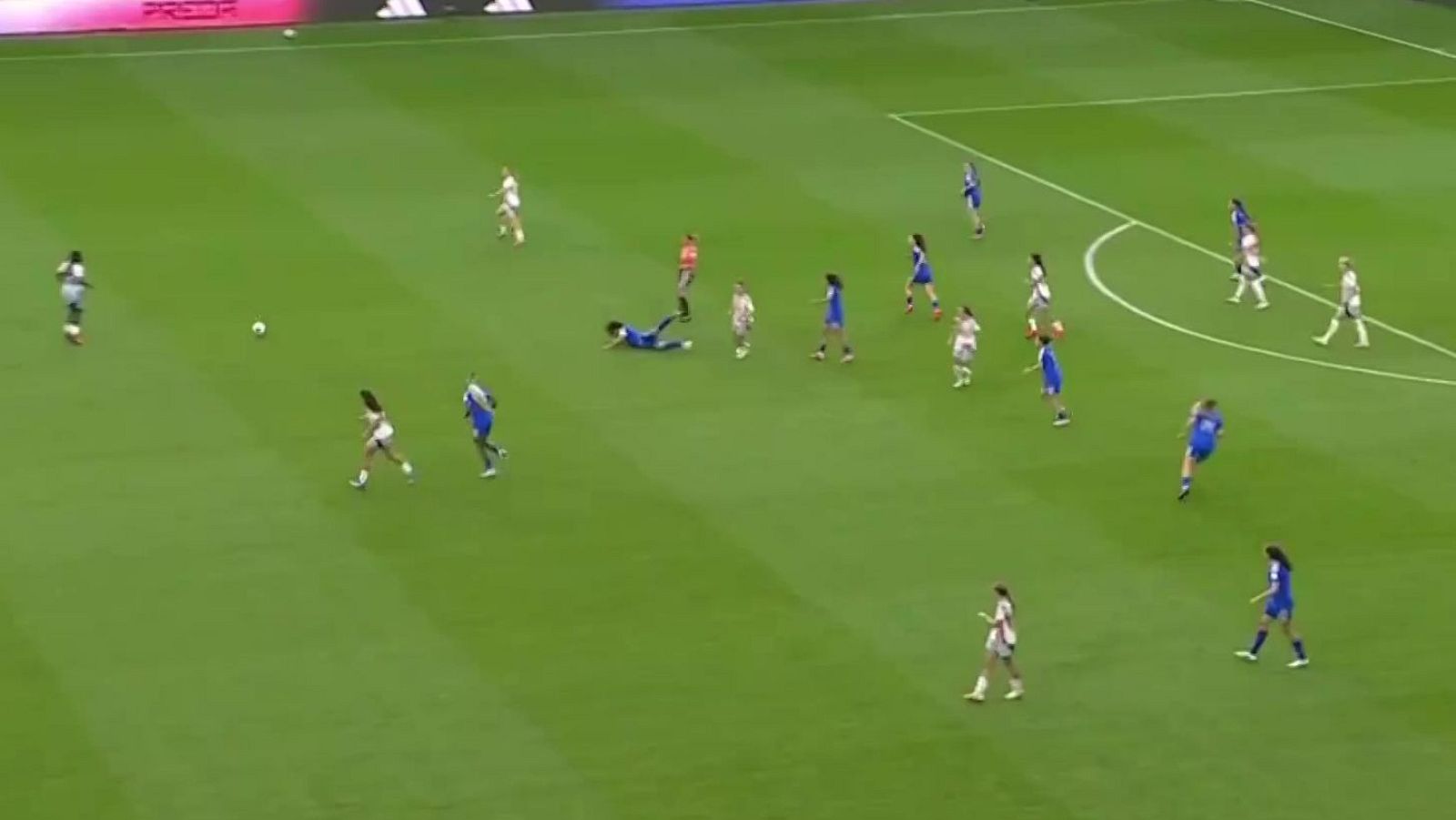 Vista aérea de un partido de fútbol femenino con jugadoras en movimiento, una en el suelo cerca del balón, en un campo de césped con líneas blancas y vallas publicitarias al fondo.