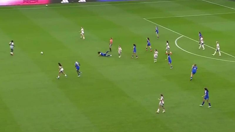 Vista a�rea de un partido de f�tbol femenino con jugadoras en movimiento, una en el suelo cerca del bal�n, en un campo de c�sped con l�neas blancas y vallas publicitarias al fondo.