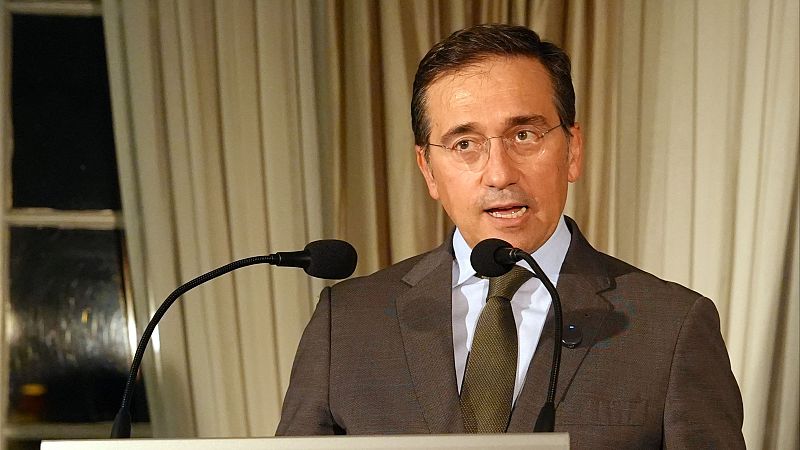 José Manuel Albares, Ministro de Asuntos Exteriores, habla en un evento. Lleva traje, gafas y corbata, con micrófonos frente a él y cortinas de fondo.