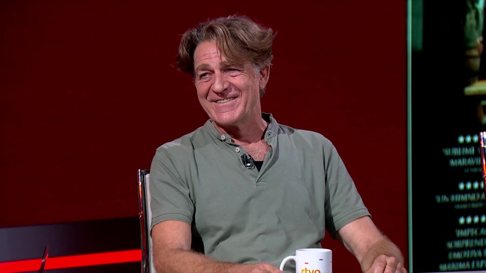 Periodista deportivo Juan Carlos Rivero sonríe en estudio de TV, con polo verde, taza RTVE y pantalla con texto positivo y estrellas.
