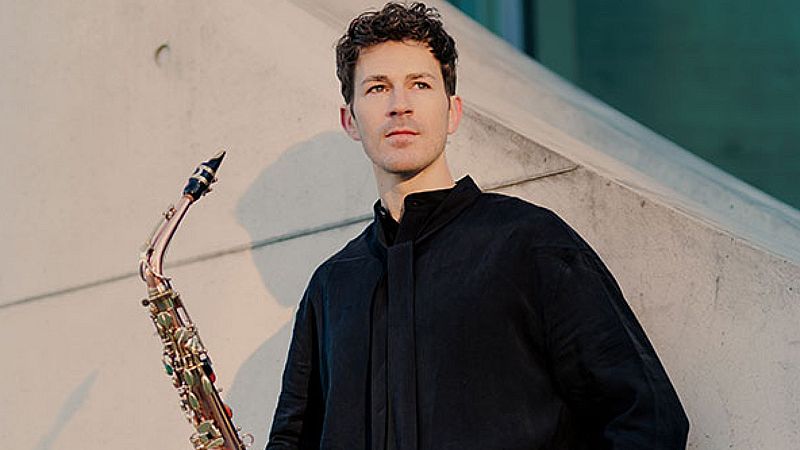 Retrato del saxofonista Carlos Giménez con su instrumento dorado y vistiendo de negro frente a una estructura de hormigón en Berlín.