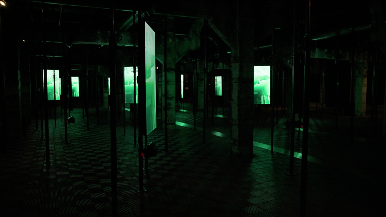 Instalación artística en sala oscura con pantallas verticales de luz verde brillante proyectando contenido abstracto. Se aprecian pilares y vigas en penumbra. CONTEXTO: Desatados.