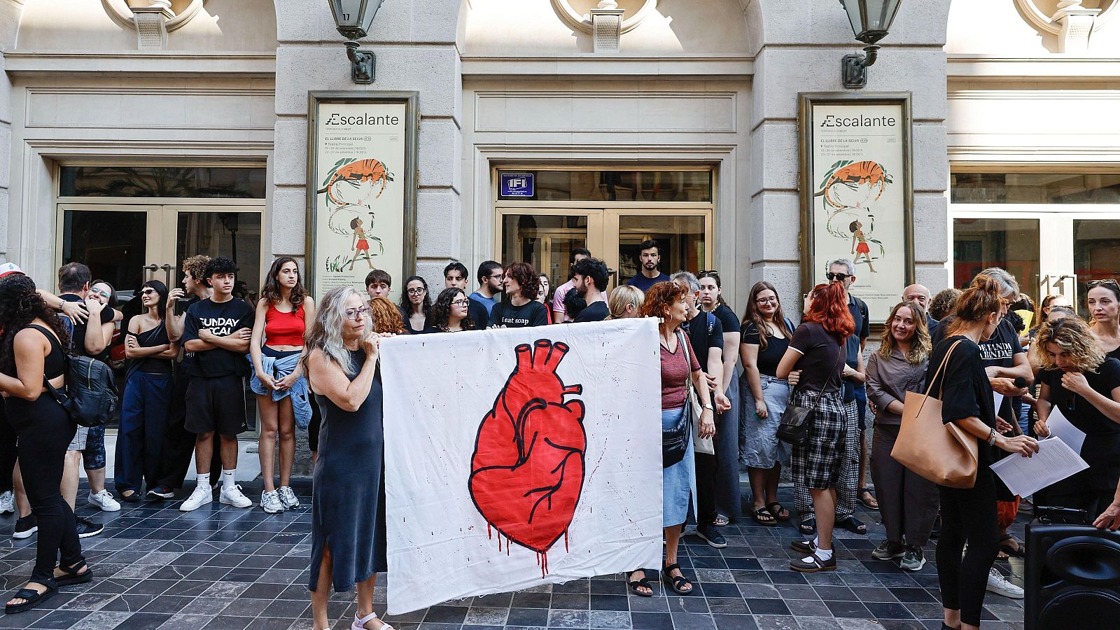 Concentración de personas frente a edificio clásico con pancarta de corazón rojo. Carteles laterales con "Escalante" e ilustraciones.