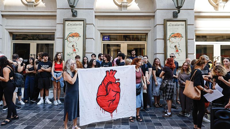 Concentración de personas frente a edificio clásico con pancarta de corazón rojo. Carteles laterales con "Escalante" e ilustraciones.