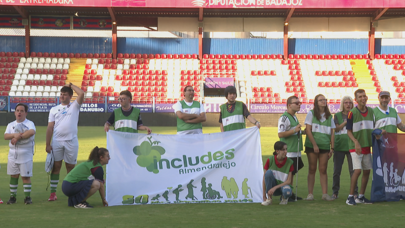 Grupo diverso en campo de fútbol con pancartas de inclusión que dicen "includes Almendralejo" y muestran siluetas de personas con capacidades diferentes.