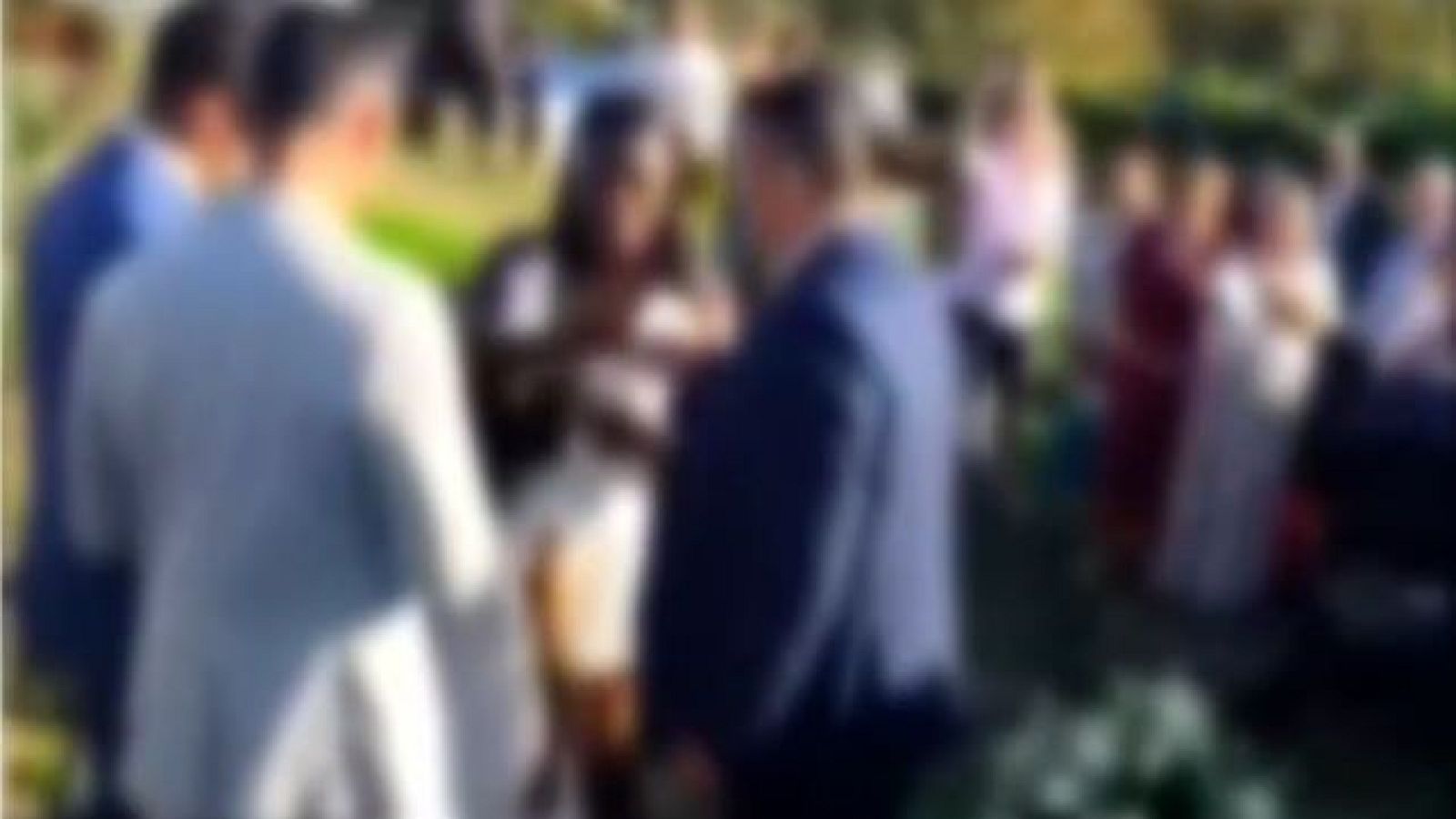 Grupo de personas reunidas al aire libre, con vestimenta formal o semi-formal, en un entorno con vegetación. La imagen está muy desenfocada, impidiendo la identificación de detalles.