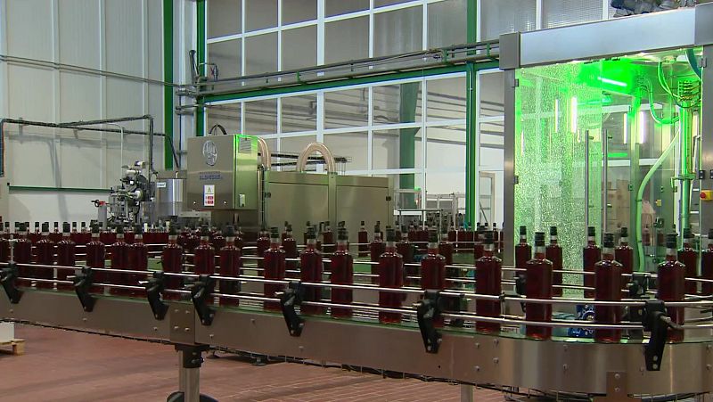 Cinta transportadora con botellas de líquido rojo oscuro en una planta de embotellado industrial, junto a maquinaria de llenado y una sección de tratamiento con luz verde.