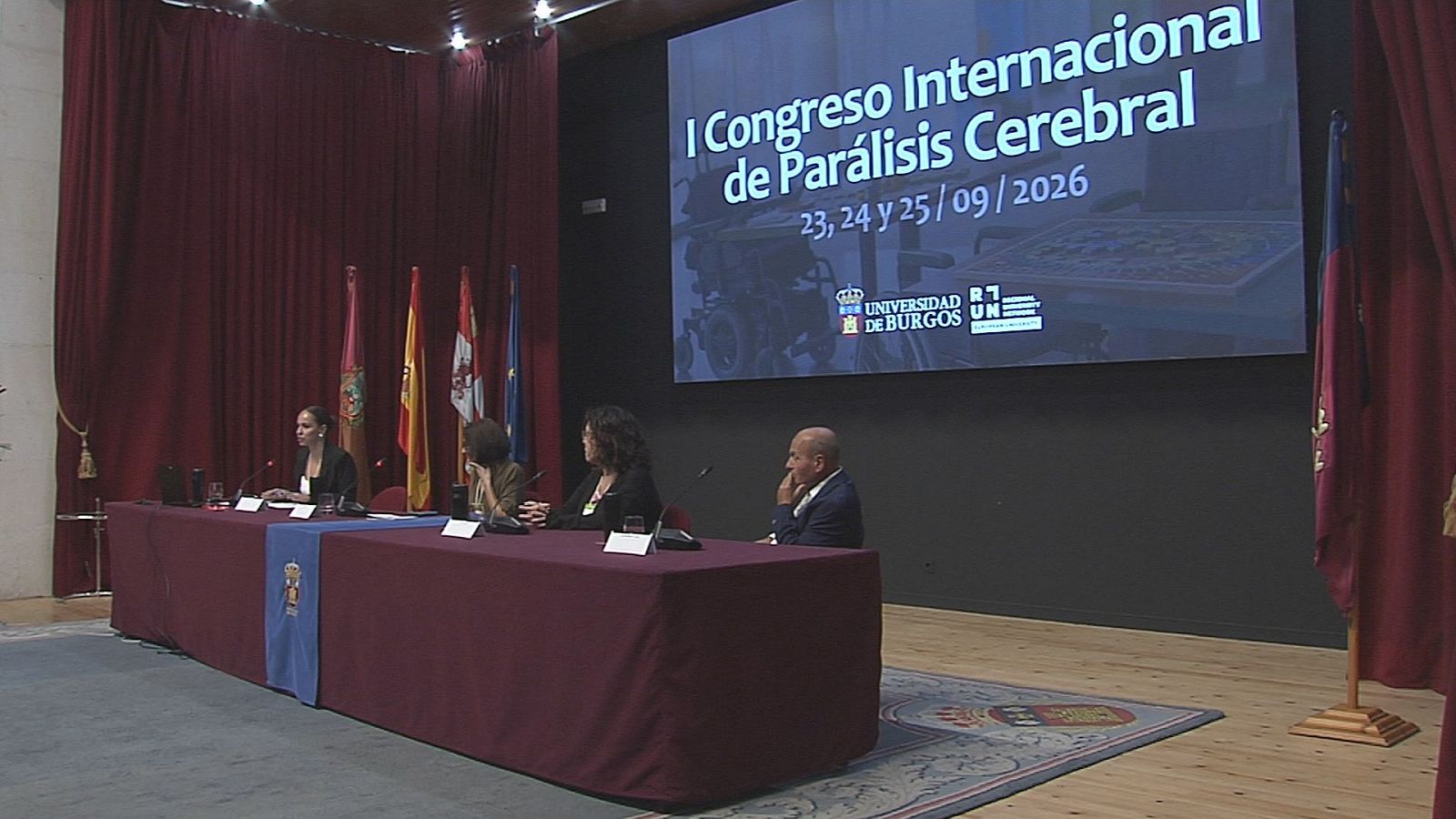 Cuatro ponentes en una mesa durante el I Congreso Internacional de Parálisis Cerebral (23-25/09/2026), organizado por la Universidad de Burgos. Banderas de España y Castilla y León presentes.
