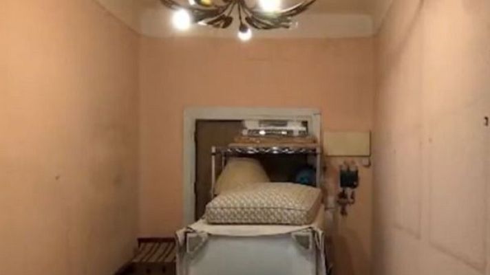 Habitación interior con paredes salmón, techo blanco y lámpara clásica. Estantería metálica con electrodoméstico y objetos. Electrodoméstico blanco cubierto con tela y cojín geométrico en primer plano.