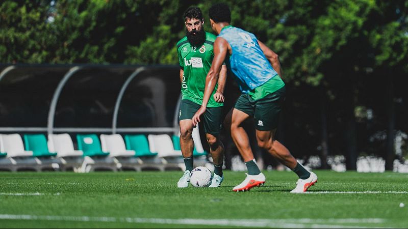 Asier Villalibre, con barba y camiseta verde número 12, entrena en un campo de césped junto a un compañero con camiseta de teñido anudado azul y blanco.