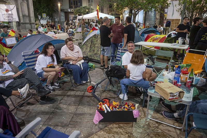 Campamento de protesta en Ceuta con personas, tiendas, banderas de España y un cartel "HAY UNA CARTA PARA TI". Se ven mesas con Lays, Coca-Cola y botellas.
