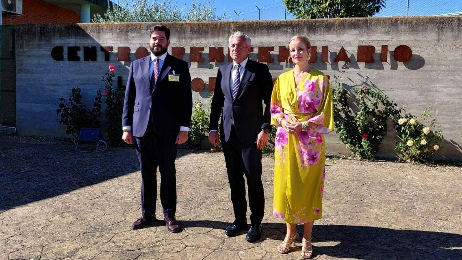 Tres personas posan frente a un muro con la inscripción "CENTRO PENITENCIARIO", adornado con rosas. Nicanor Sen y Marta Tuoya, junto a un hombre con traje, participan en un acto institucional.