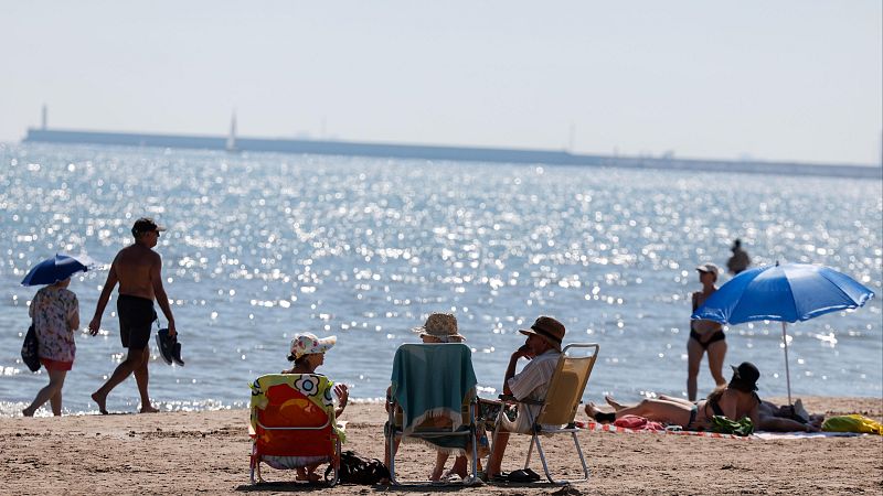 Playa soleada con gente disfrutando del mar y la arena. Una pareja pasea, otros descansan en sillas o bajo sombrillas, y alguien se refresca en el agua.
