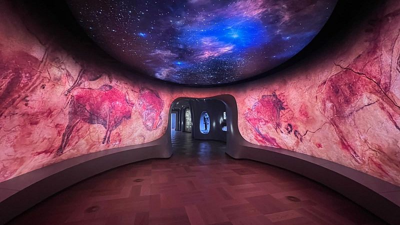 Pasillo moderno con arte rupestre de bisontes en paredes curvas y cielo estrellado proyectado en el techo. Suelo de parquet en espiga. Vitrinas iluminadas al fondo.