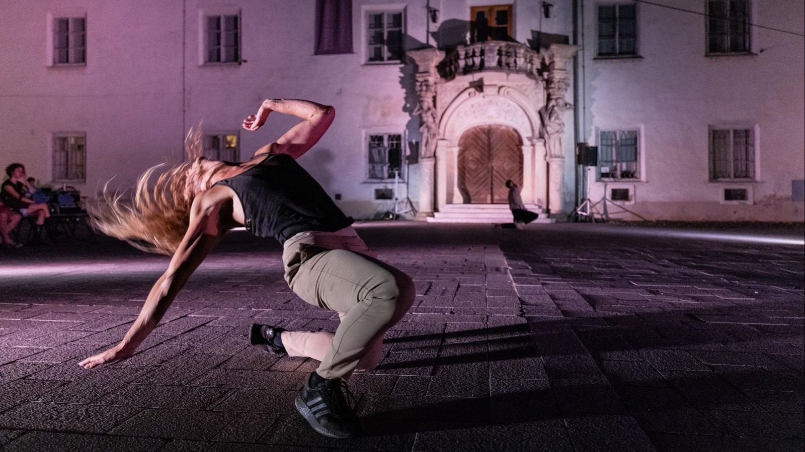 Mujer atl�tica en pose de danza en plaza urbana con edificio hist�rico iluminado. Luz p�rpura y haz blanco resaltan movimiento y energ�a.
