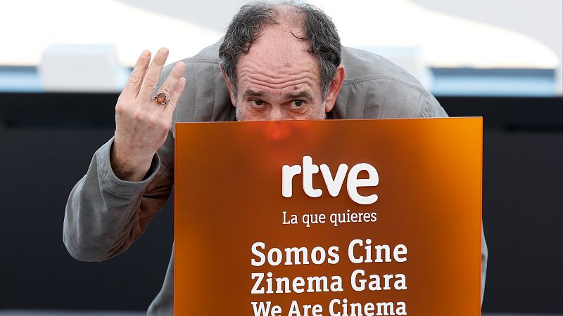 Carlos Saura, director de cine español, posa frente a un cartel de RTVE con el lema "Somos Cine". Su mano izquierda, con un anillo, se alza mientras mira intensamente.