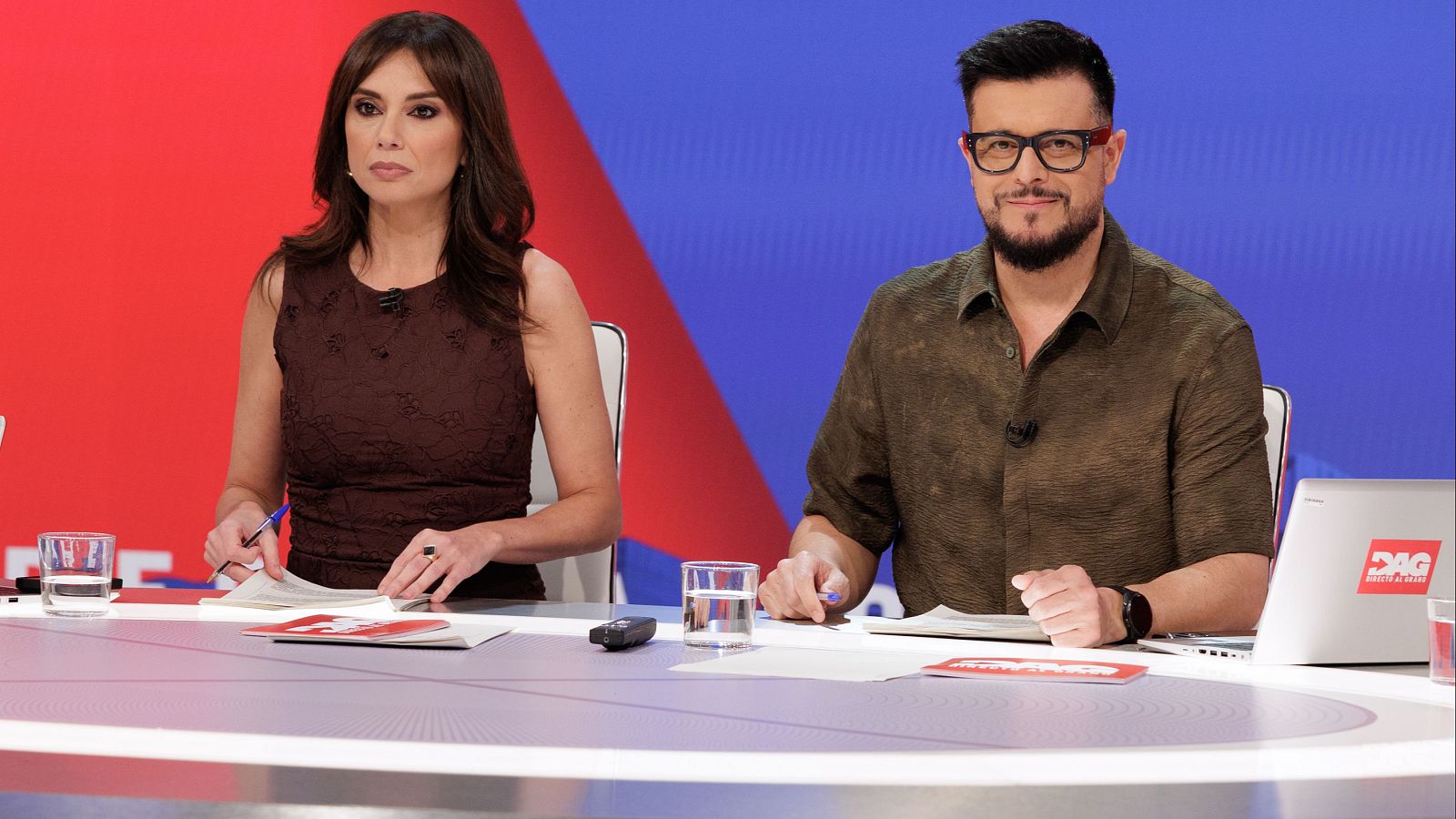 Ana Rosa Quintana y Joaquín Prat en un estudio de televisión, sentados ante una mesa con un portátil que muestra "DIRECTO AL GRANO". Fondo rojo y azul.
