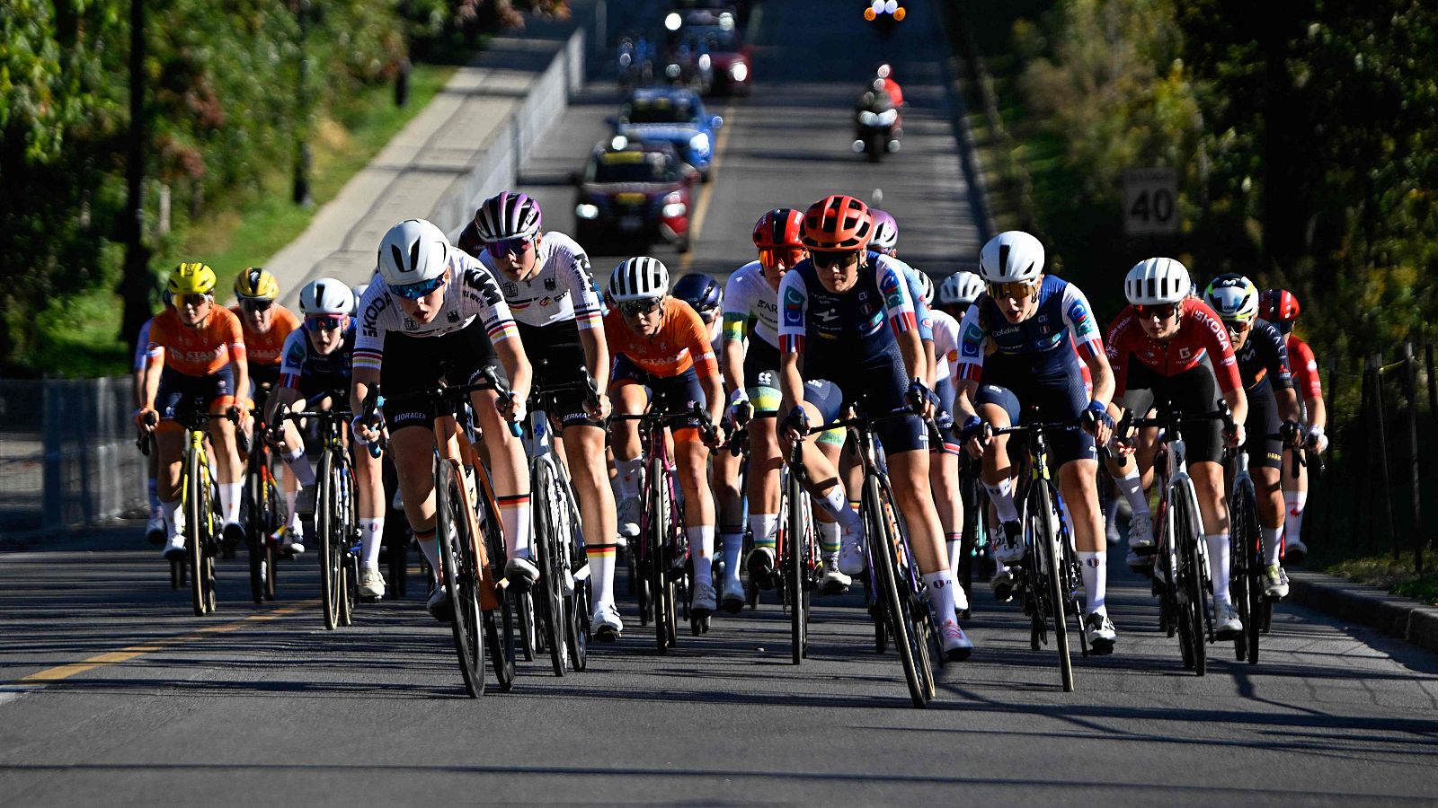 Ciclistas profesionales en plena ascensión por carretera asfaltada, con vegetación a los lados y señal de límite de 40 km/h. Vehículos de apoyo siguen la carrera.