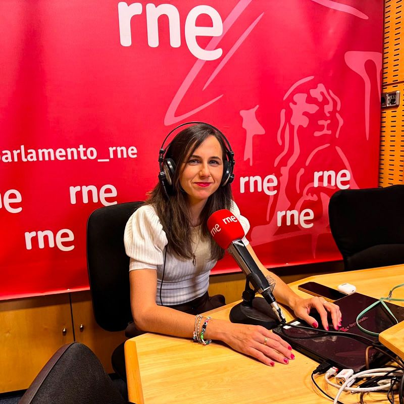 Ione Belarra sonríe a cámara en un estudio de radio, con auriculares y pulseras. Detrás, el logo "rne" destaca sobre un fondo rojo.
