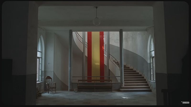 Interior de un vestíbulo antiguo con una gran bandera de España en el centro, escaleras a ambos lados y ventanas arqueadas. Luz tenue y atmósfera solemne.