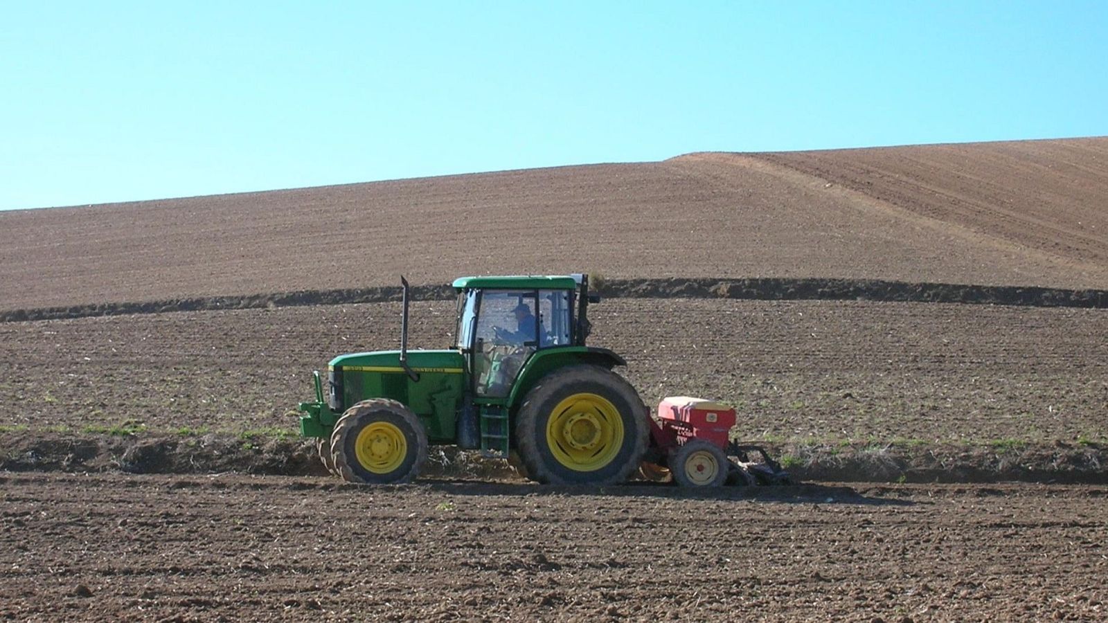 Tractor verde con apero rojo labrando un campo de tierra en Aragón, bajo un cielo azul despejado. La imagen captura la actividad agrícola mecanizada.