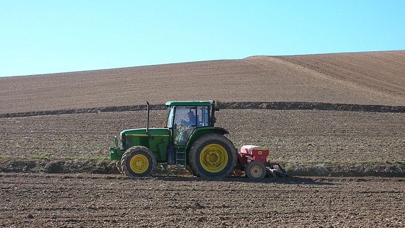 Tractor verde con apero rojo labrando un campo de tierra en Aragón, bajo un cielo azul despejado. La imagen captura la actividad agrícola mecanizada.