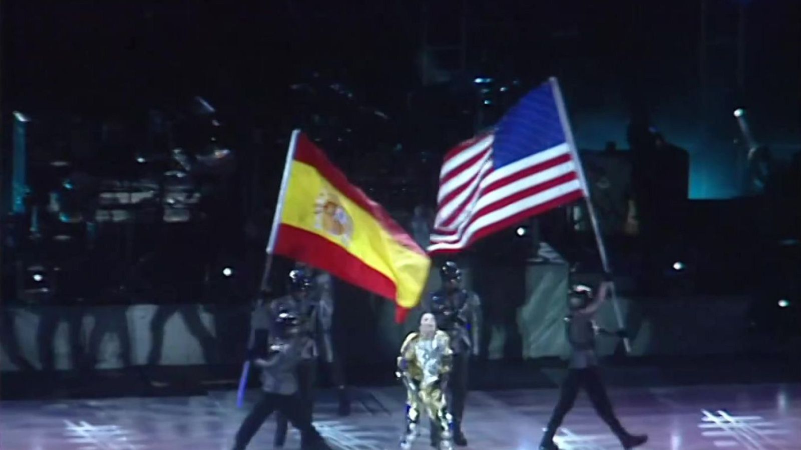 Michael Jackson en un traje brillante, flanqueado por figuras con cascos que portan las banderas de España y EE.UU. en un escenario con luces.