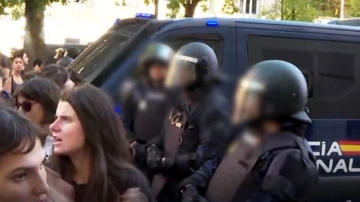 Agentes antidisturbios de la Policía Nacional en formación frente a un furgón policial, mientras un grupo de personas observa la escena en un entorno al aire libre.