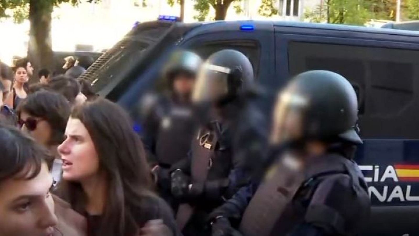 Agentes antidisturbios de la Policía Nacional en formación frente a un furgón policial, mientras un grupo de personas observa la escena en un entorno al aire libre.